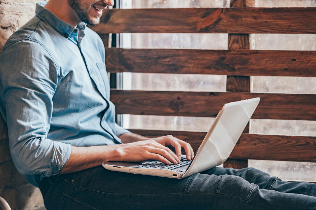 Do blogs still help with SEO in 2023? A side view close-up of handsome young man working on laptop and smiling whilesitting at windowsill